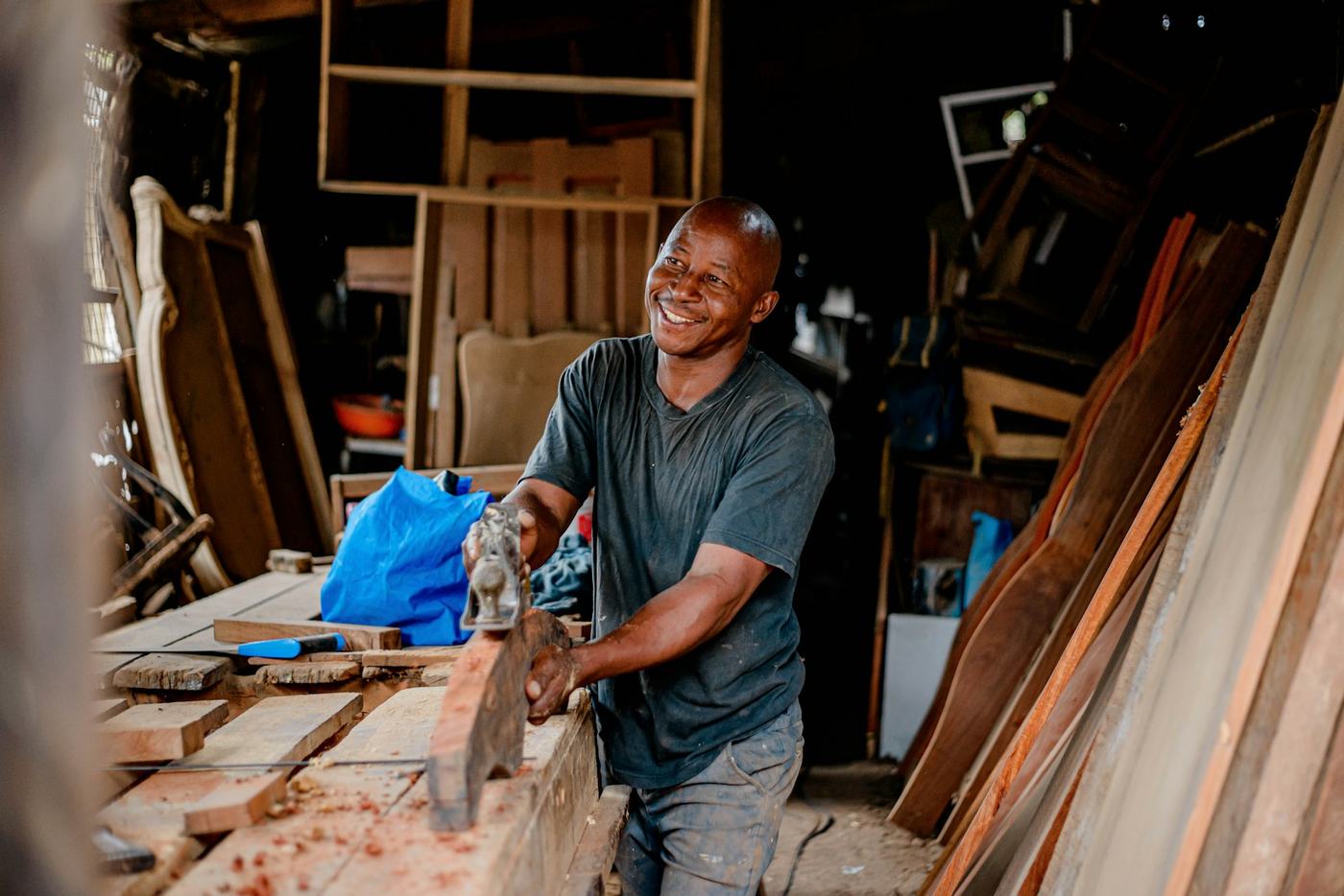 Carpenter smiling while working on furniture surrounded by wood and reclaimed pieces